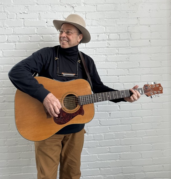 Charlie Maguire, wearing a brown cap and striped scarf, plays guitar in a room lined with hanging acoustic guitars.