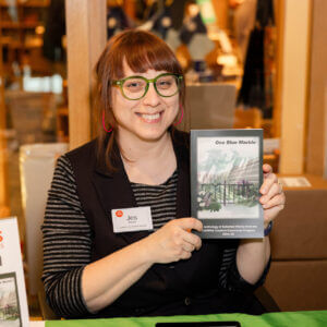 A smiling woman, Jes Reyes holding the COMPAS Anthology of Student Writing.