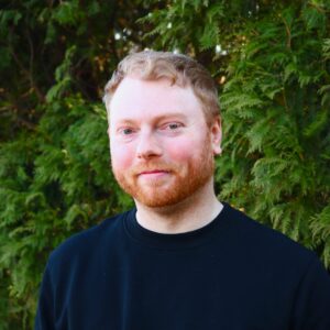 Headshot of Jacob Snyder Timmons, a man with short light red hair and a beard, wearing a black shirt, standing in front of green foliage.