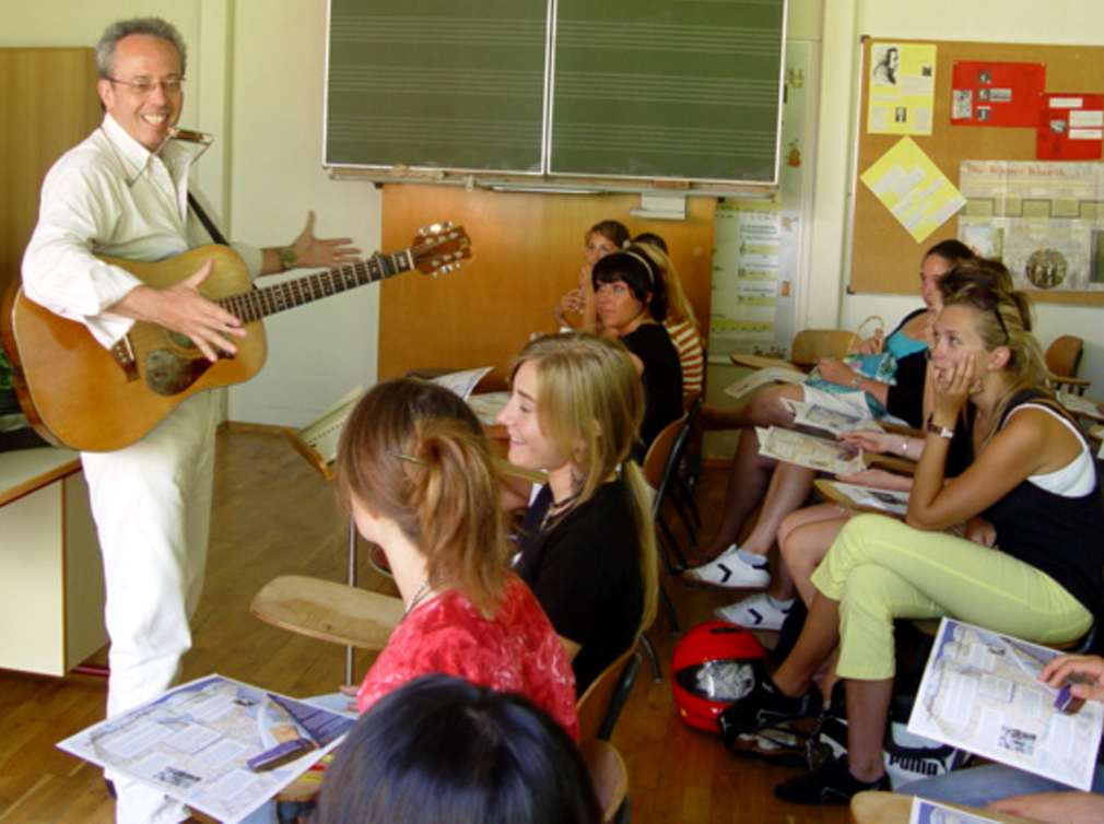 Photo of COMPAS teaching artist Charlie Maguire standing in front of a classroom of middle school students with his guitar while he offers a lesson.