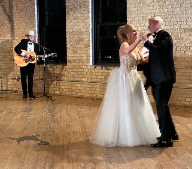 Caitlin Raasch and her father dancing a father-daughter dance at Caitlin's wedding. She is wearing a white wedding dress, and her father is wearing a black tuxedo. In the background, Charlie can be seen playing his guitar for them to dance to. He is wearing a black tuxedo and a white cowboy hat.