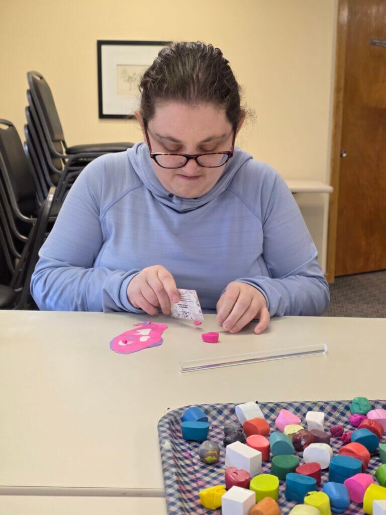 A person wearing glasses and a light blue hoodie sits at a table, focused on shaping a small piece of bright pink clay with a card. In front of them is a tray filled with colorful clay pieces, and a flattened pink form rests on the table nearby. Stacked chairs and a framed picture are visible in the background.
