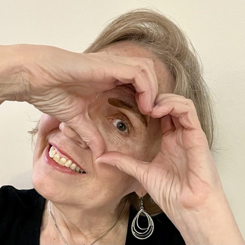 A smiling person holds both hands around one eye, forming a heart shape with their fingers.