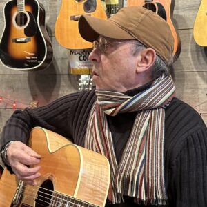Charlie Maguire, wearing a brown cap and striped scarf, plays guitar in a room lined with hanging acoustic guitars.