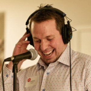 Smiling man wearing headphones and a checkered shirt speaks into a microphone. He appears to be enjoying himself, possibly hosting or recording audio. A name tag reading 'Andrew' is visible on his chest."