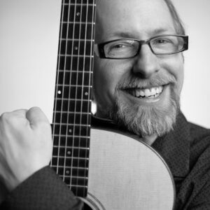 Black and white image of Scott Keever holding a guitar close and smiling.