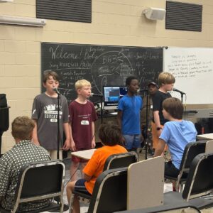 Several teen boys standing at the front of the classroom, a blackboard behind them, microphones in front of them, performing for classmates, who are sitting in rows facing them.