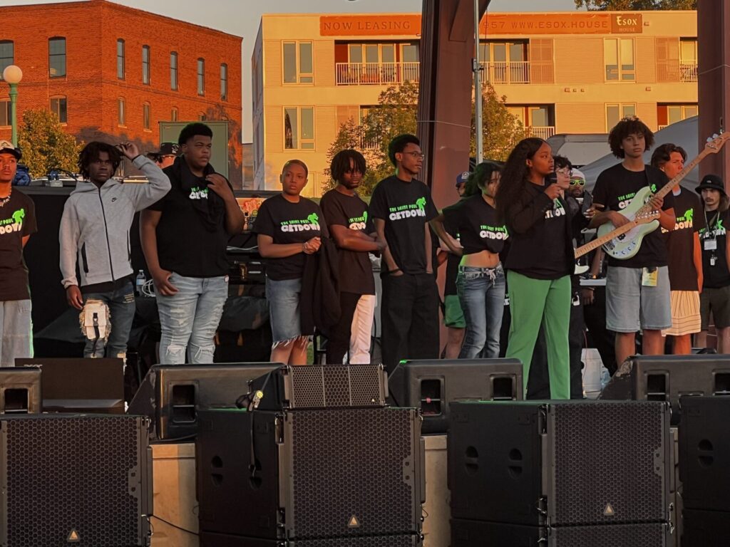 A group of young performers stand together on an outdoor stage at sunset, wearing matching black T-shirts with text on the front, as one person speaks into a microphone. Large speakers are set up in front of the stage, with buildings visible in the background.