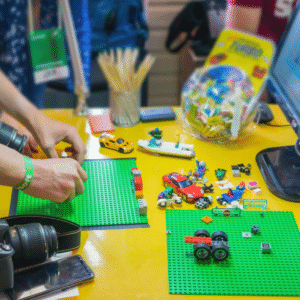 Hands arranging LEGO pieces and small toy vehicles on green baseplates at a yellow table, with building materials spread out, suggesting a hands-on creative or stop-motion animation activity.