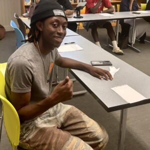 Simmia sitting at a table working on a piece of writing giving a thumbs up gesture.