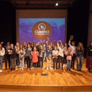 A diverse group of elementary and middle school students stand on a stage holding books and raising their fists in celebration, with a large screen behind them displaying the COMPAS “Making Creators” logo.