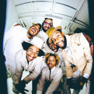 Joe Davis and four other members of The Poetic Diaspora look up at the camera in a joyful group shot, wearing coordinated neutral tones and smiling widely in a bright white space