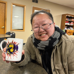 Smiling person wearing glasses and a scarf sits at a table, holding a white mug decorated with a colorful abstract drawing.”