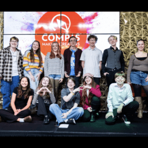 Twelve middle and high school student poets pose together on stage following the COMPAS My Voice. My Time! Youth Spoken Word Showcase, smiling in front of a large COMPAS display.
