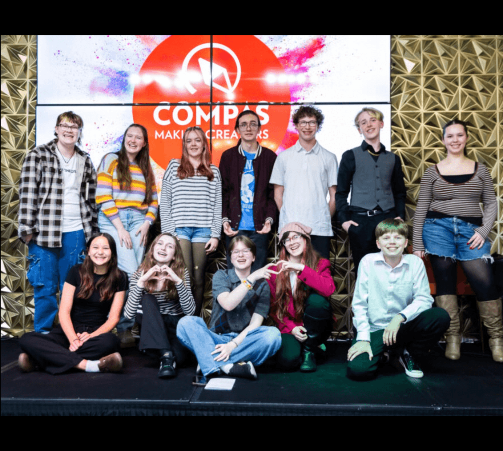Twelve middle and high school student poets pose together on stage following the COMPAS My Voice. My Time! Youth Spoken Word Showcase, smiling in front of a large COMPAS display.