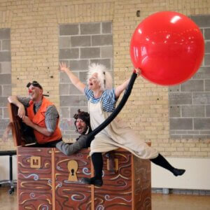 Three artists of Cahoot!?, two with clown noses, in movement with a big red ball.